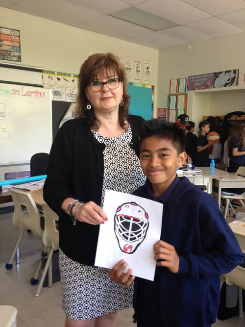 Catherine Rondina stands in a classroom. A student in a blue shirt is standing next to her. He is holding a hand drawn picture of a goalie's mask.